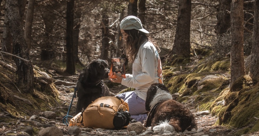 2 dogs and their owner resting during a hike. The owner is kneeling between the dogs and feeding the Oven-Baked Tradition