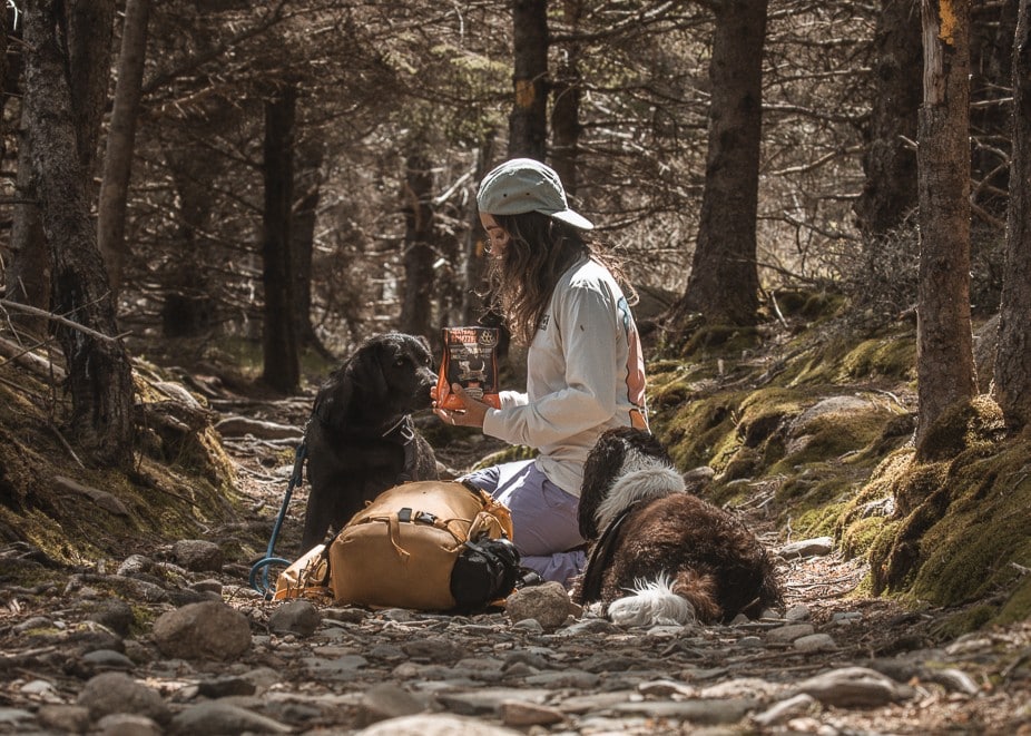 2 dogs and their owner resting during a hike. The owner is kneeling between the dogs and feeding the Oven-Baked Tradition